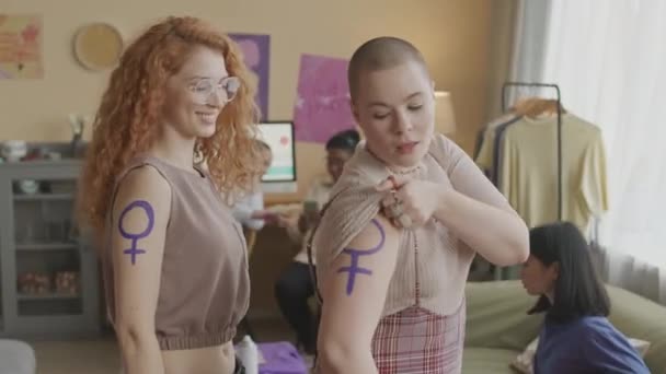 Средний портрет двух молодых кавказских активисток, демонстрирующих фиолетовый феминистский символ на своих плечах во время общинной встречи с другими женщинами дома - Кадры, видео
