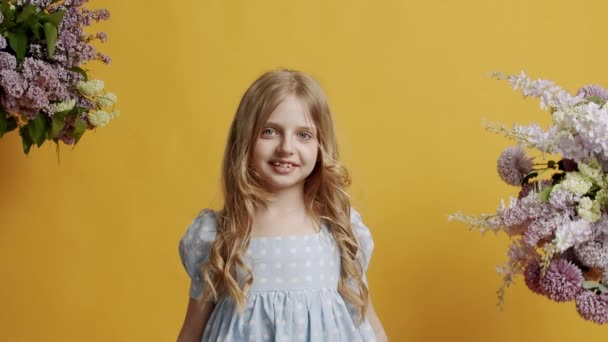 7-vuotias pikkutyttö on pukeutunut vaaleansiniseen mekkoon ja näyttää kauniisti huoneeseen keltaisella taustalla studiossa. Kaunis pieni tyttö katsoo kameraan ja hymyilee ystävällisesti. - Materiaali, video