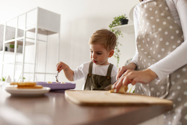 mladá matka v zástěře dává sušenky a ovesnou kaši svému synovi k jídlu v kuchyni doma, 2-letý chlapec snídá se svým rodičem, žena krájí banán a krmí dítě - Fotografie, Obrázek