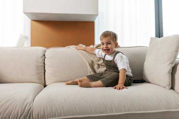 mały chłopiec 2 lata siedzi na miękkiej wygodnej kanapie w domu i śmieje się, szczęśliwe dziecko uśmiecha - Zdjęcie, obraz
