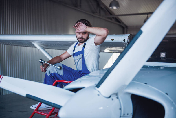 Guapo barbudo mecánico de uniforme se limpia la frente mientras reparan un avión en el hangar
 - Foto, imagen
