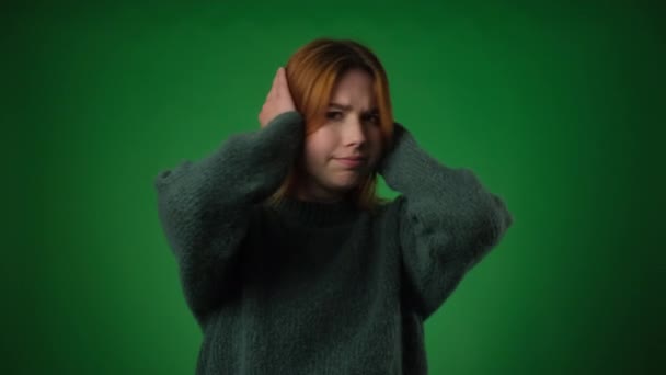 Melancholic Caucasian woman, dressed casually, covers ears with hands on a green backdrop. Her sorrowful expressions and the gesture of shielding ears convey a desire to block out unwanted sounds. - Footage, Video