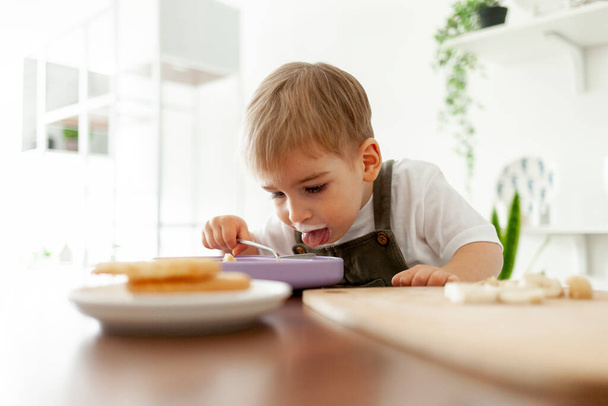 zabawny mały chłopiec 2 lata je śniadanie sam w kuchni z nabiału, dziecko jest brudne z jedzeniem, je jogurt z łyżką i uśmiecha się - Zdjęcie, obraz