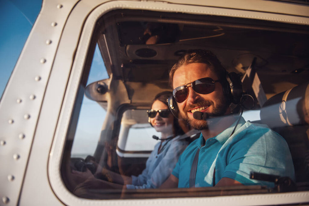 Beautiful couple in aviation headsets is looking  at camera and smiling while sitting in aircraft ready to fly - Photo, Image