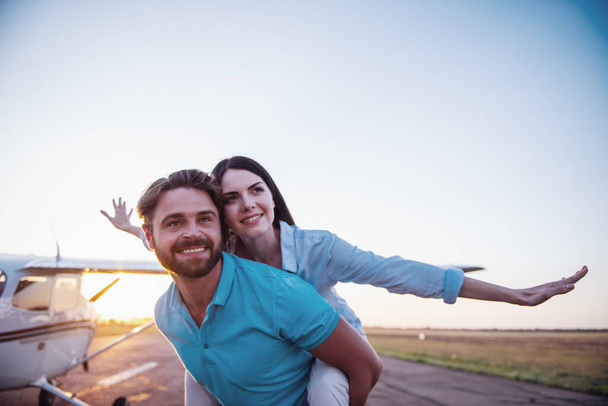 schönes romantisches Paar schaut weg und lächelt, während es auf dem Startplatz in der Nähe des Flugzeugs steht, Mädchen huckepack - Foto, Bild
