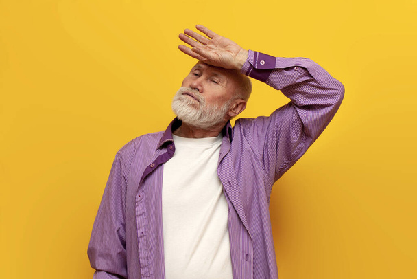 tired old bald grandfather in a purple shirt wipes sweat from his forehead on yellow isolated background, sad elderly pensioner sweating m shows hot temperature - Photo, Image