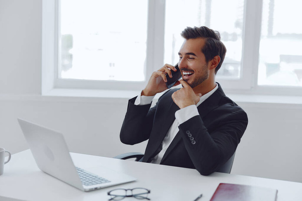 Мужчина сидит менеджер счастливый бизнес-ноутбук Интернет мобильный телефон молодой успех говорить белый костюм работает успешный портрет компьютерный бизнесмен офис привлекательная улыбка - Фото, изображение