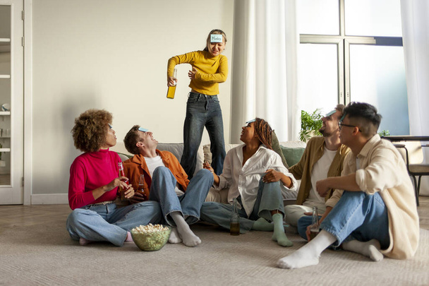 multiracial group of young people sitting at home with beer and popcorn and playing charades and having fun with friends, Asian girl showing and pretending to joke - Photo, Image