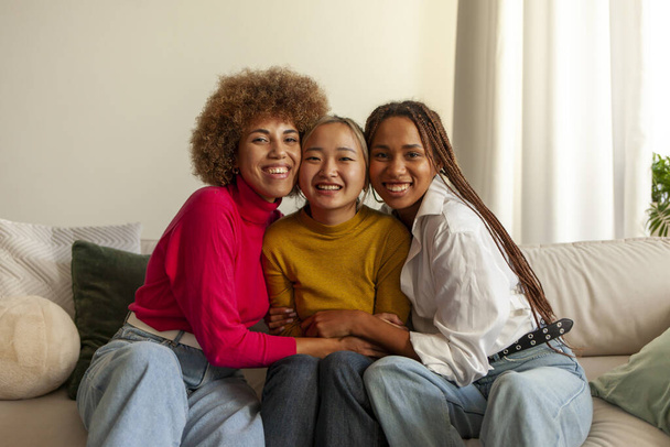 Afro-Amerikaanse vrouwen en Aziatische vrouw zitten op de bank thuis en knuffelen, interraciale vriendinnen glimlachen en kijken naar de camera - Foto, afbeelding