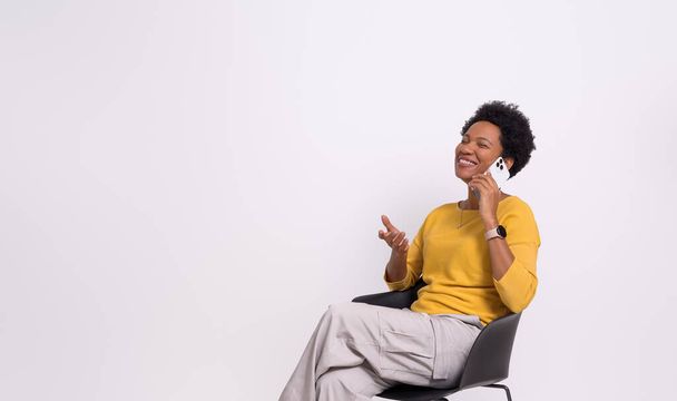 Femme d'affaires souriante avec coiffure afro parler sur téléphone intelligent sur chaise sur fond blanc - Photo, image