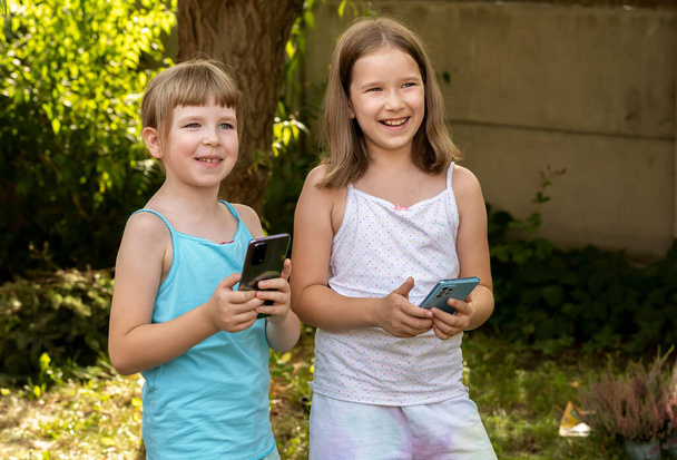 Happy school leeftijd kinderen twee jonge meisjes kinderen houden hun mobiele telefoons in de buitenlucht, buiten glimlachend. Met behulp van moderne technologie, apps, games, sms abstract concept, echte mensen - Foto, afbeelding