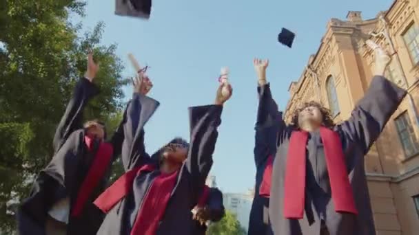 Vista ad angolo basso di scapoli multiculturali che celebrano la loro laurea e lanciano cappelli in aria - Filmati, video