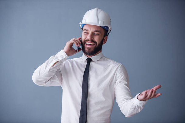 Handsome young bearded businessman in suit and protective helmet is talking on the mobile phone and smiling, on gray background - Photo, Image