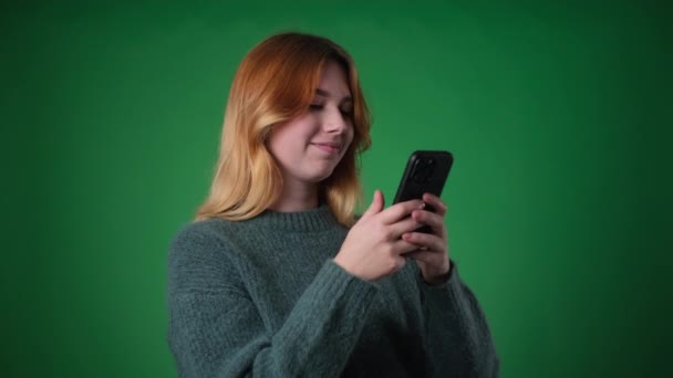 Cheerful Caucasian woman, dressed casually, smiles while using phone against a vibrant green backdrop. Her radiant expression and engaged demeanor convey a sense of joy and connection. - Footage, Video