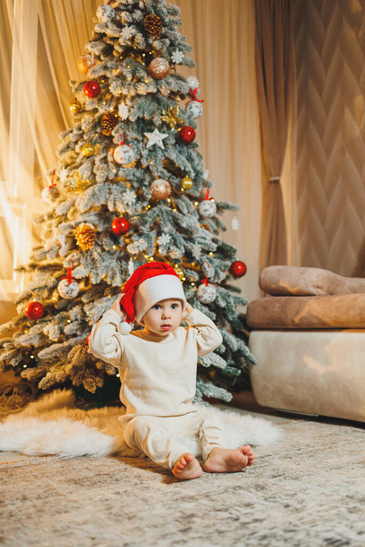 Netter kleiner Junge mit Weihnachtsmütze und Weihnachtsbaum. Kinderneujahr. Weihnachtsbaum. Weihnachtsstimmung zu Hause. - Foto, Bild