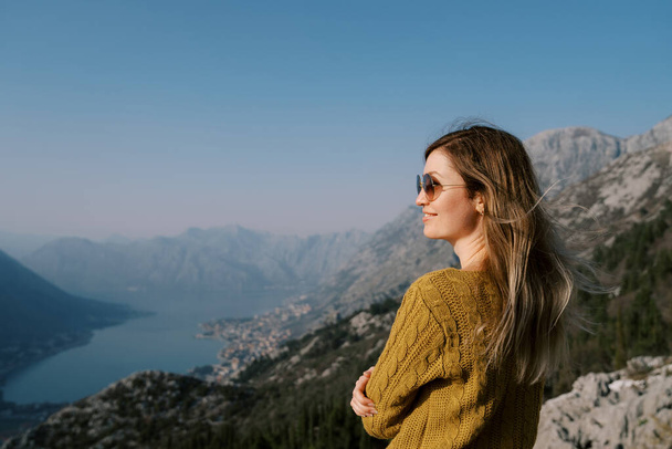 Усміхнена дівчина в сонцезахисних окулярах стоїть на горі над затокою Котор. Чорногорія. Вид збоку. Високоякісна фотографія - Фото, зображення