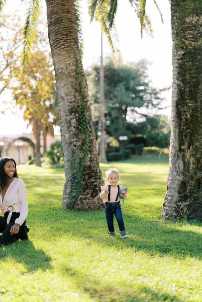 Little laughing girl with a teddy bear walks on a sunny lawn past a smiling woman. High quality photo - Photo, Image