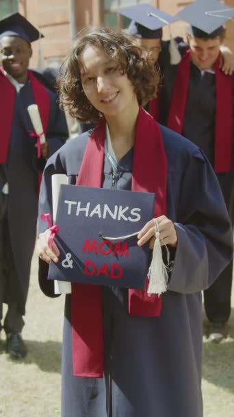 Ritratto verticale di giovane donna che tiene il cappello con gratitudine alla madre e al padre mentre si diploma all'università, guardando la macchina fotografica - Filmati, video