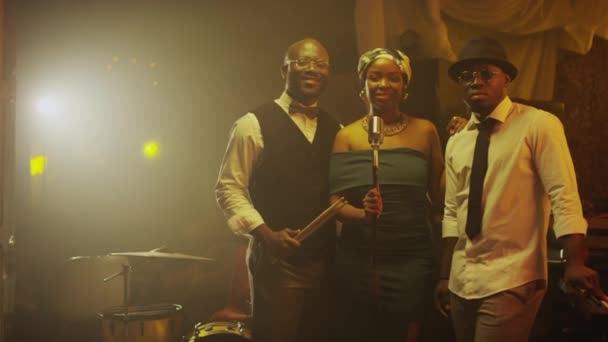 Middelgroot portret van hedendaags Afro-Amerikaans jazztrio met mooie zangeres die glimlacht naar de camera op het podium met warm gedimd licht in chique jazzclub - Video
