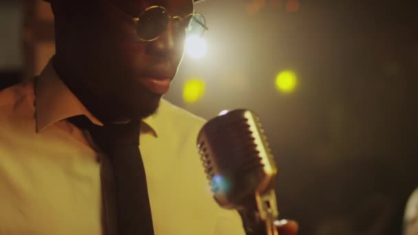 Nuori komea musta mies mustissa pyöreissä aurinkolaseissa, valkoisessa paidassa ja mustassa solmiossa laulaa bluesia retro-mikrofonissa klubin konsertin aikana yöllä - Materiaali, video