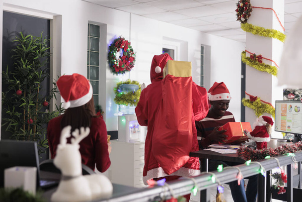 Счастливые работники удивляются Санта-Клаусу подарками на Рождество. Коллега, переодетый в рождественский костюм, дарит подарки улыбающимся коллегам в празднично украшенном офисе - Фото, изображение