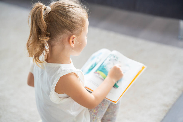 Söpö pikkutyttö istuu ja selailee kirjaa, jossa on kuvia saduista. Laadukas kuva - Valokuva, kuva