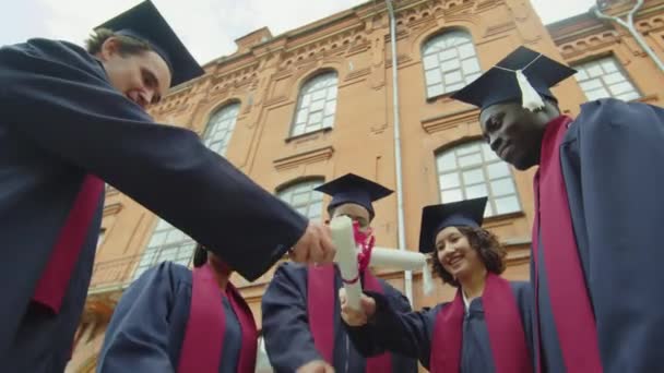 Vista ad angolo basso di studenti in abiti e cappelli alzare i loro diplomi dopo la cerimonia di laurea al di fuori dell'università - Filmati, video