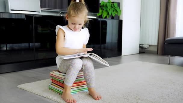 La petite fille est assise sur une pile de livres pour enfants et feuillette un livre avec des contes de fées. Images 4k de haute qualité - Séquence, vidéo