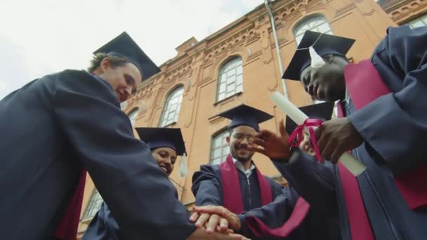 Vista a basso angolo di gruppo di diversi studenti laureati eccitati che fanno gesto di squadra e alzano le mani vicino all'università - Filmati, video