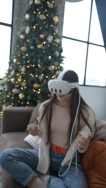 Egy fiatal nő játszik egy játékot egy virtuális valóság headset, ül egy karácsonyfa mellett egy kanapén. Kiváló minőségű 4k felvételek - Felvétel, videó