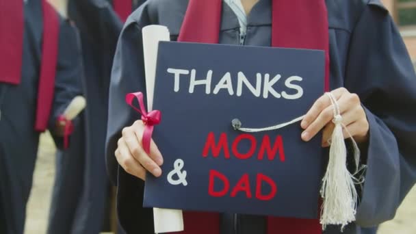 Colpo ritagliato di studentessa irriconoscibile che tiene il cappello universitario con testo ringrazia mamma e papà durante la cerimonia di laurea - Filmati, video