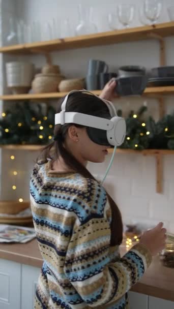 Una joven con un atuendo de moda cocina con un auricular de realidad virtual en una cocina con temas navideños. Imágenes de alta calidad 4k - Imágenes, Vídeo