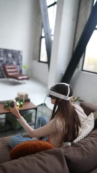 Een jonge vrouw, in de buurt van een kerstboom, houdt van gamen terwijl ze een virtual reality-headset gebruikt die op een bank zit. Hoge kwaliteit 4k beeldmateriaal - Video