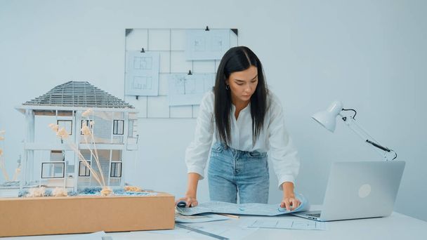 Young beautiful caucasian architect puts blueprint on the table and compare with house model while inspect hos model construction carefully at modern office with blueprint striped behind. Immaculate. - Photo, Image