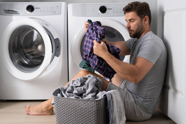 Man with clothes near washing machine. Handsome man sits in front of washing machine. Loads washer with dirty laundry. Man cleaning clothes. Housework for single alone guy. Home laundry - Photo, image