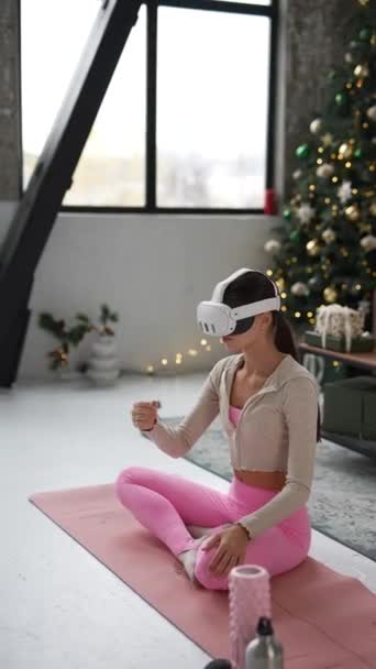 Zaangażowany w jogę w domu podczas świąt Bożego Narodzenia, szczupła młoda kobieta wykorzystuje zestaw słuchawkowy wirtualnej rzeczywistości. Wysokiej jakości materiał 4k - Materiał filmowy, wideo