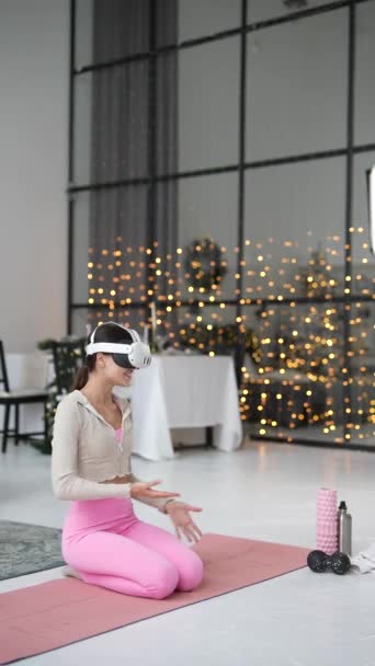 Mit einem Virtual-Reality-Headset nimmt eine schöne junge Frau in der Nähe eines Weihnachtsbaums Yoga-Posen ein. Hochwertiges 4k Filmmaterial - Filmmaterial, Video