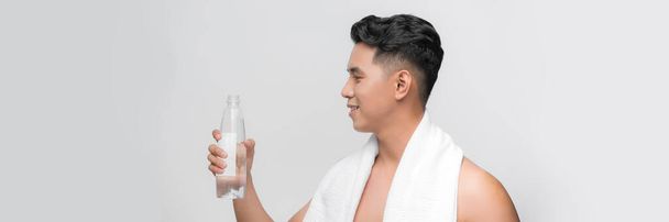 Jovem confiante com toalha nos ombros segurando garrafa com água e sorrindo
 - Foto, Imagem