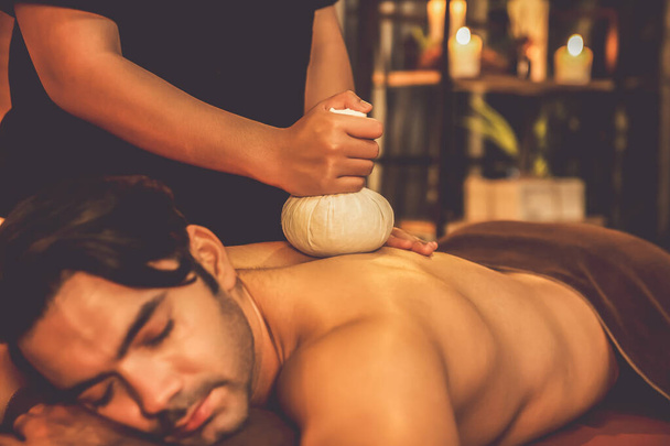 Gorące ziołowe kulkowe spa masaż ciała leczenie, masażysta delikatnie kompresuje worek ziołowy na ciele człowieka. Spokojny i spokojny aromaterapii rekreacji w ciepłym zapaleniu świec w salonie spa. Cichy zapach - Zdjęcie, obraz