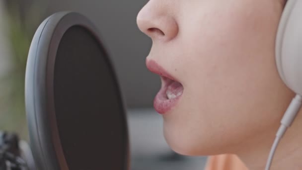 Κοντινό πλάνο της νεαρής κοπέλας με ακουστικά που τραγουδάει στο μικρόφωνο με τα μάτια κλειστά ενώ ηχογραφεί μουσική στο στούντιο του σπιτιού - Πλάνα, βίντεο