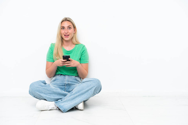 Jovem caucasiana sentada no chão isolada em fundo branco surpreendida e enviando uma mensagem - Foto, Imagem