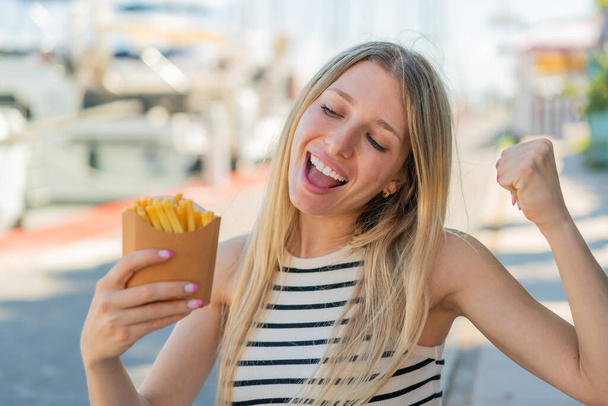 Mladá blondýna žena drží smažené chipsy venku slaví vítězství - Fotografie, Obrázek