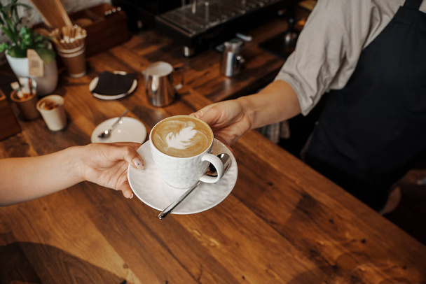 Barista ojentaa kauniisti muotoillun cappuccinon lattella asiakkaalle puisessa kahvilassa - Valokuva, kuva