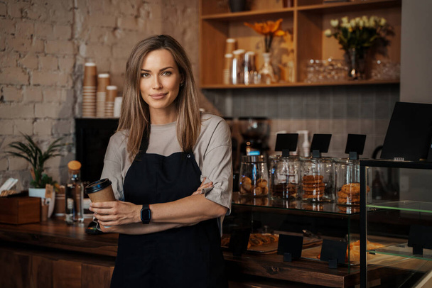 A portrait of a poised female barista holding a takeaway coffee cup, standing in her modern cafe - Photo, Image