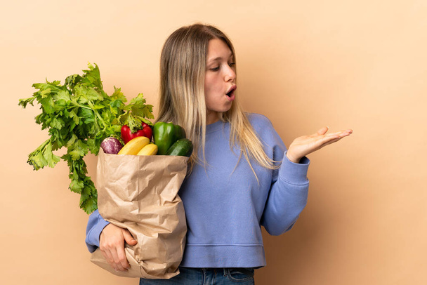 Young blonde woman holding a bag full of vegetables over isolated background holding copyspace imaginary on the palm - Photo, Image