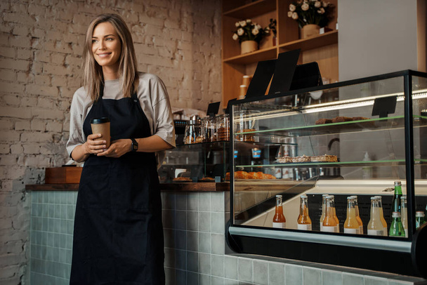Een portret van een vrouwelijke barista die een meeneemkoffiebeker vasthoudt en in haar moderne café staat - Foto, afbeelding