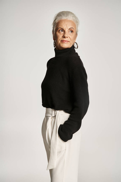счастливая деловая женщина средних лет в элегантной одежде позирует с руками в карманах на сером фоне - Фото, изображение