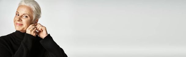 szczęśliwy w średnim wieku kobieta biznesu w eleganckim stroju nosić kolczyki obręczy na szarym tle, baner - Zdjęcie, obraz