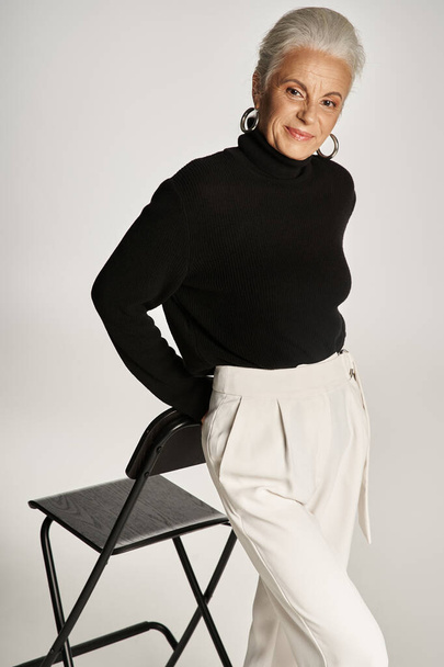 mujer de negocios de mediana edad en traje casual inteligente y pendientes de aro de pie cerca de la silla en gris - Foto, imagen