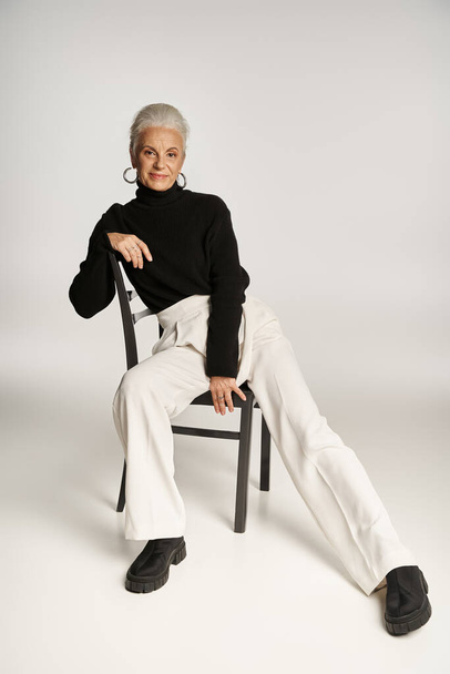 affascinante donna di mezza età in elegante abbigliamento casual e orecchini a cerchio seduto sulla sedia su grigio - Foto, immagini